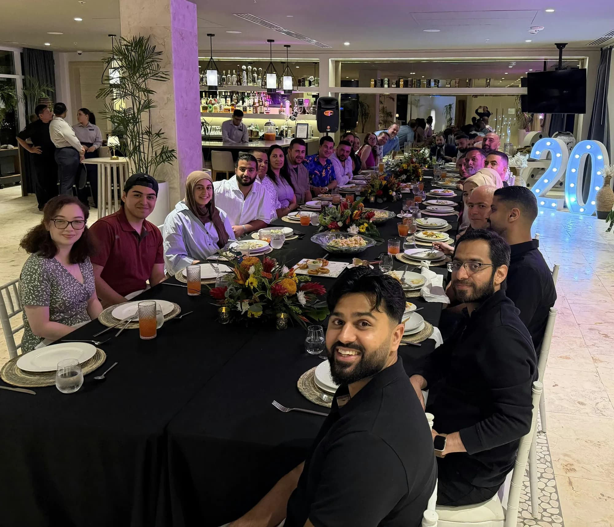Guests gathered around a long dinner table, with a lit-up number 20 sign in the background