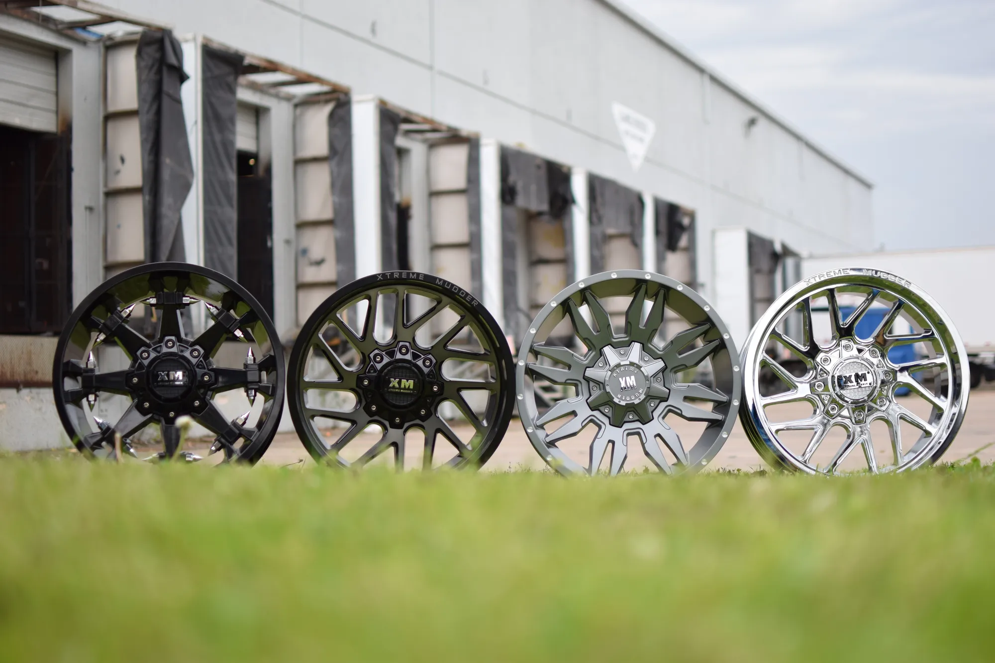 Four XM wheels in black and chrome finishes standing on the grass in front of the warehouse loading docks